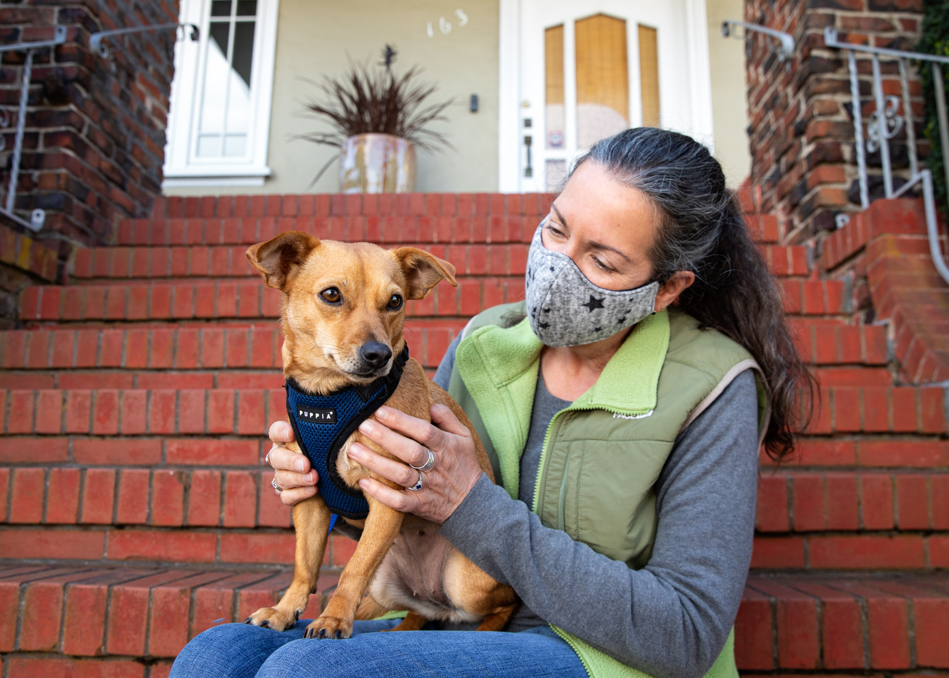 middle-aged woman wearing face mask and holding a chihuahua mix dog on front steps of her house